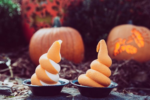 Two soft serve ice cream cones, one orange and white swirl, the other solid orange, with carved pumpkins in the background.