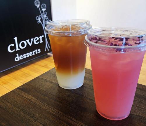 Two colorful drinks in clear cups sit on a wooden table, with a sign for "clover desserts" in the background.