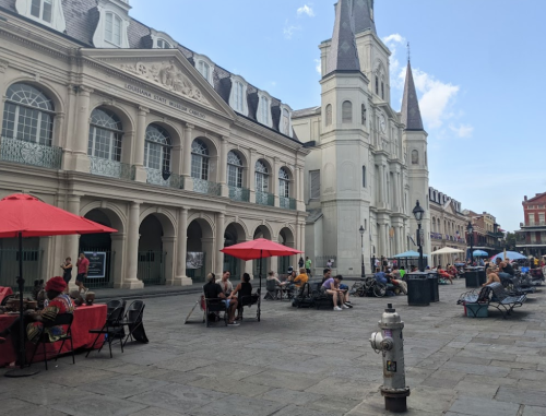 A lively square with people dining under red umbrellas, historic buildings, and a fire hydrant in the foreground.