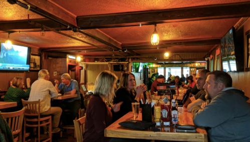A lively pub interior with patrons at tables, enjoying drinks and conversation, and TVs showing sports in the background.