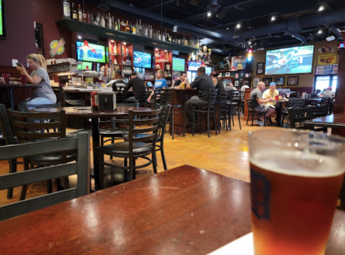 A lively bar scene with patrons at the counter, TVs showing sports, and a glass of beer in the foreground.