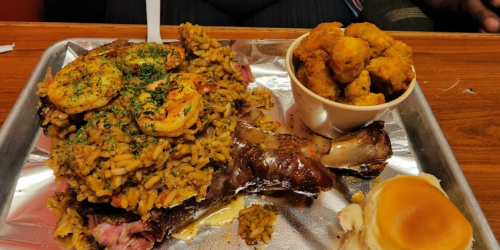 A large platter featuring seasoned rice topped with shrimp, a smoked meat leg, and a side of fried food.