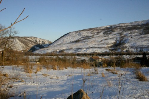Snow-covered hills and a valley under a clear blue sky, with sparse vegetation and distant houses visible.