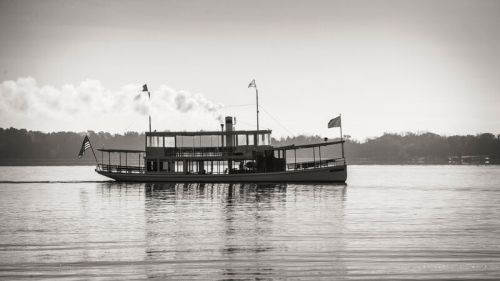 A vintage steamboat glides across a calm lake, with smoke billowing from its stack and flags waving in the breeze.