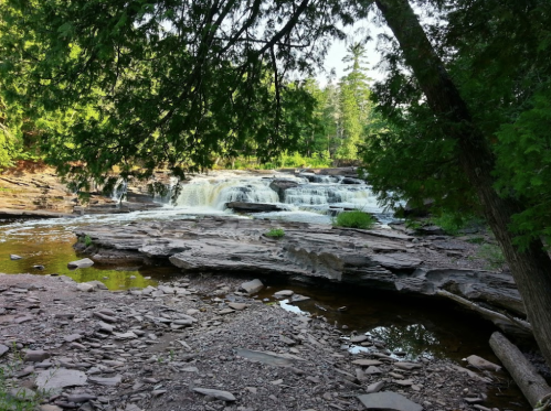 A serene waterfall cascades over rocky terrain, surrounded by lush green trees and a calm river.