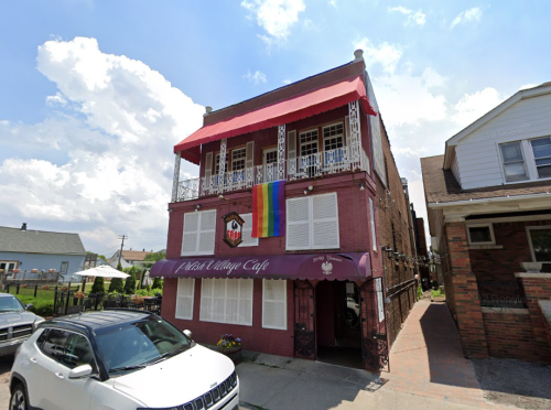 A colorful café with a rainbow flag hanging from the balcony, set against a bright blue sky and fluffy clouds.