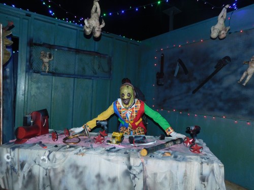 A creepy figure in a colorful outfit stands at a cluttered table with tools and eerie dolls in a dimly lit room.