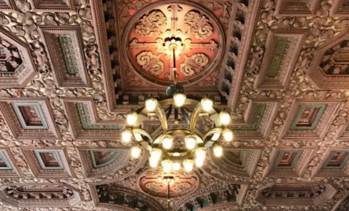 Ornate ceiling with intricate designs and a vintage chandelier illuminated by warm lights.
