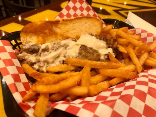 A basket with a cheesesteak sandwich and crispy French fries, served on a checkered paper liner.