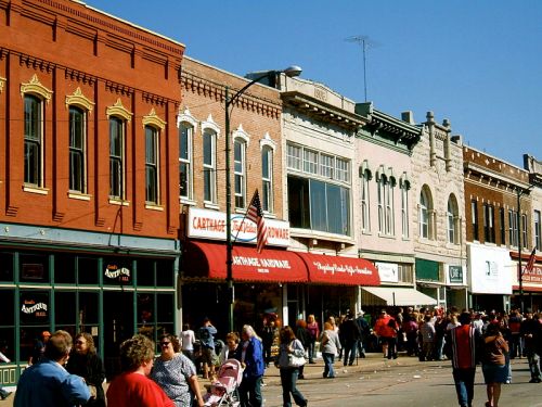 A bustling street scene with historic buildings, shops, and a crowd of people enjoying a sunny day.
