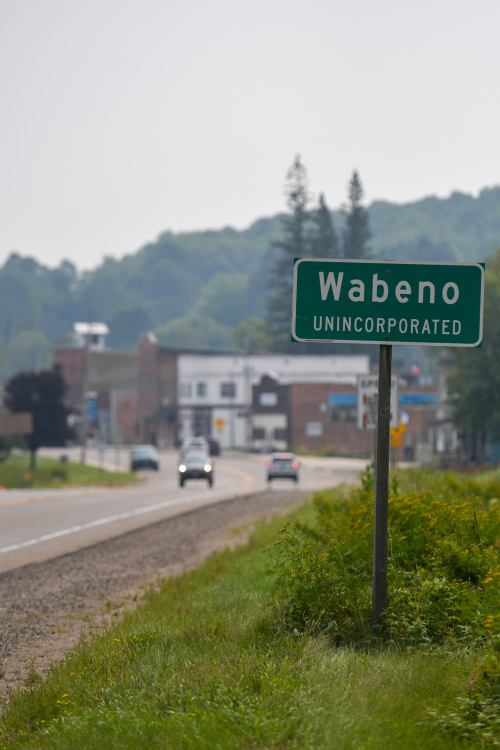 Sign for Wabeno, an unincorporated community, with a view of the road and buildings in the background.