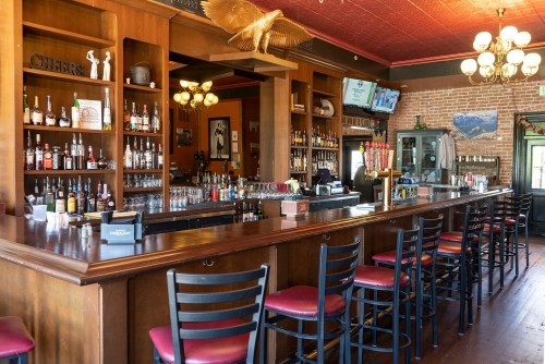 A wooden bar with high stools, shelves of liquor, and a warm, inviting atmosphere in a rustic pub setting.