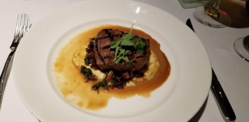 A plated dish featuring grilled meat on creamy mashed potatoes, garnished with greens and a savory sauce.
