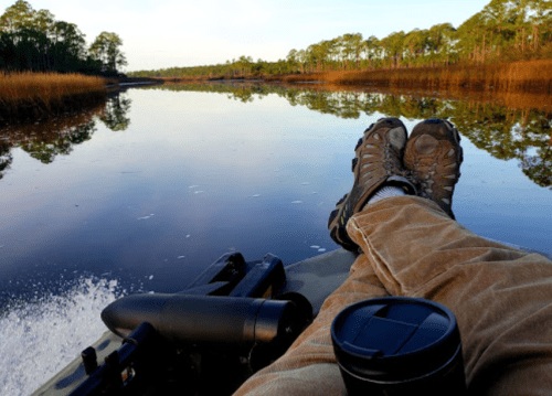 A person relaxes in a boat with feet up, surrounded by calm water and trees, holding a coffee cup.