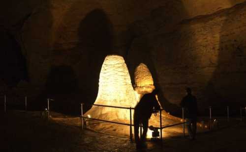 Two silhouettes explore illuminated rock formations in a dark cave, casting shadows on the walls.