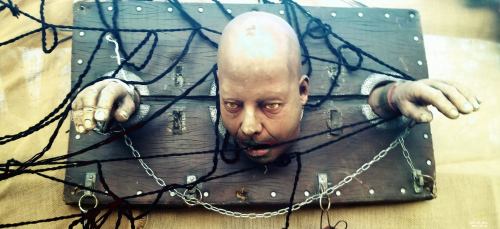 A lifelike head and hands emerge from a wooden board, entangled in ropes and chains, creating a striking visual effect.