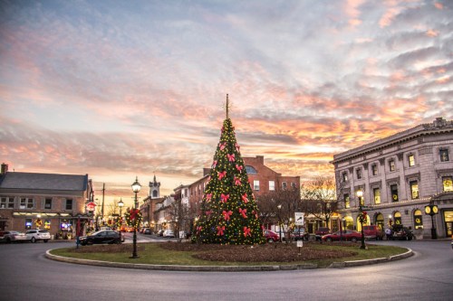 A festive Christmas tree adorned with lights and decorations stands in a town square at sunset, surrounded by buildings.