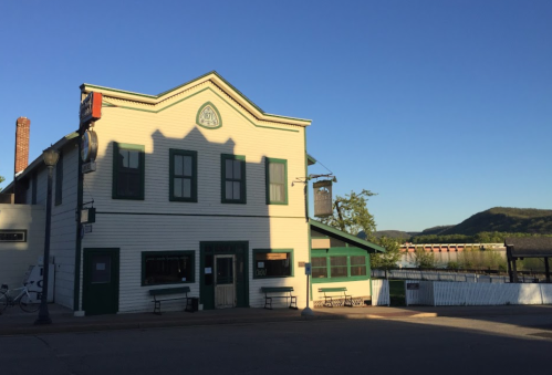 A historic two-story building with green trim, set against a clear blue sky and mountains in the background.
