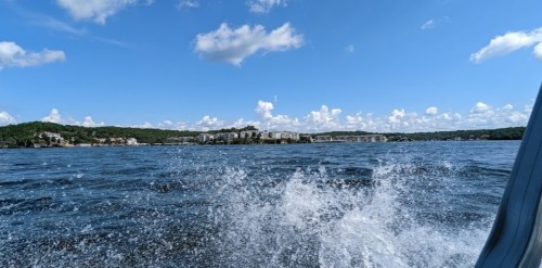 A scenic view of a lake with rippling water, blue sky, and fluffy clouds, framed by green hills and distant buildings.