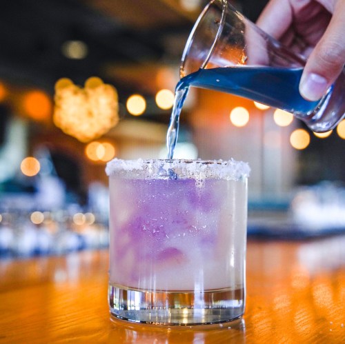 A hand pours blue liquid into a glass with a salted rim, revealing a colorful drink with bubbles and ice.