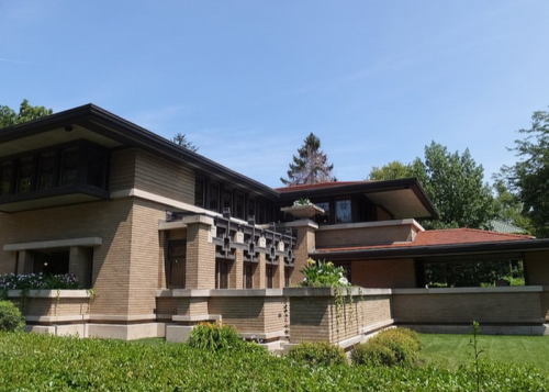 A modernist brick house with large windows, surrounded by greenery and trees under a clear blue sky.