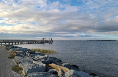A calm waterfront scene with a wooden pier, rocky shoreline, and cloudy sky reflecting on the water.