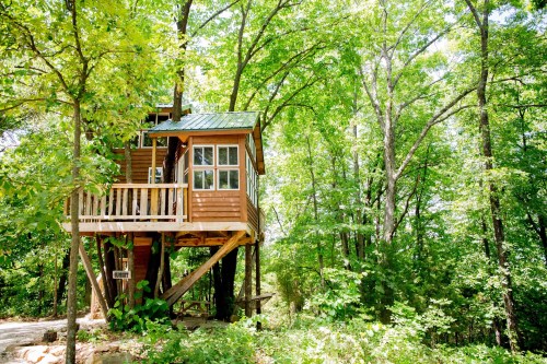 A wooden treehouse elevated among lush green trees, featuring a balcony and large windows.