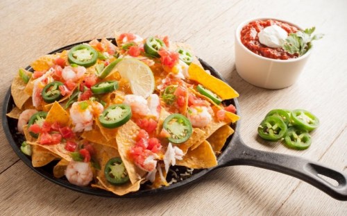 A skillet of nachos topped with shrimp, jalapeños, tomatoes, and lime, served with a side of salsa and sour cream.