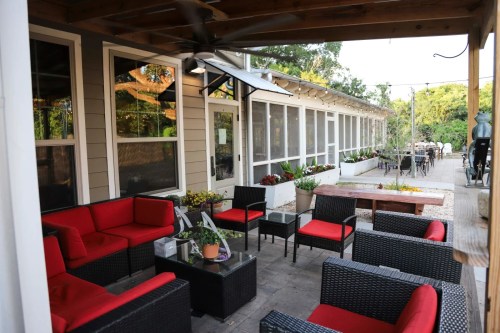 Outdoor seating area with red cushions, surrounded by greenery and a dining space in the background.