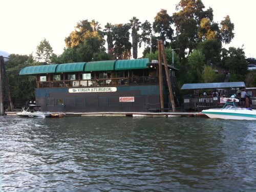 A waterfront restaurant with a green roof, surrounded by trees, and boats docked nearby on a calm river.