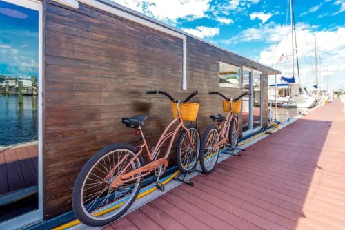 Two vintage bicycles with baskets parked beside a wooden houseboat on a sunny marina dock.