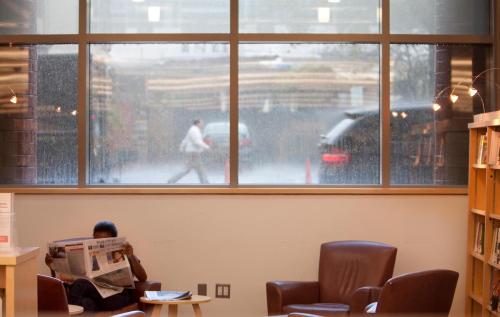 A person reading a newspaper indoors, with rain visible on the window and a blurred figure walking outside.