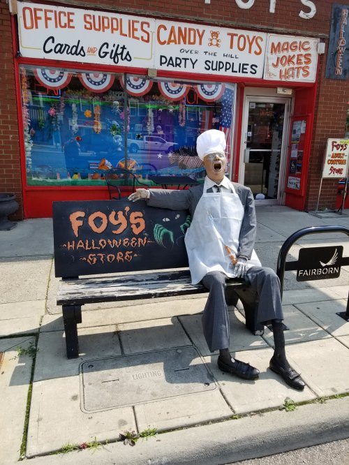 A mannequin in a chef's outfit sits on a bench outside a Halloween store, with colorful decorations in the background.
