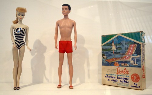 A vintage Barbie doll in a swimsuit stands next to a Ken doll in red shorts, with a Barbie furniture kit box in the background.