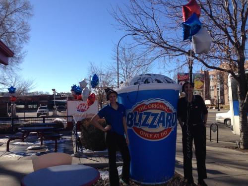 Two employees stand beside a large Blizzard cup outside a Dairy Queen, decorated with balloons and a clear blue sky.