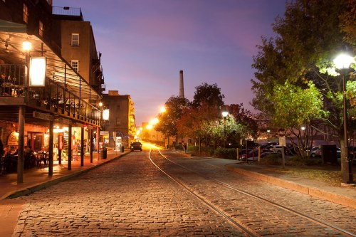 A cobblestone street at dusk, lined with trees and illuminated by street lamps, with buildings and a faint skyline in the background.