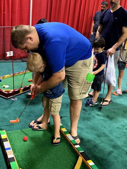 A father helps his young son practice mini-golf at an indoor event, with other families in the background.