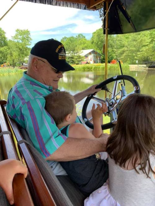 An older man steers a boat with two young children beside him, enjoying a sunny day on a calm lake.