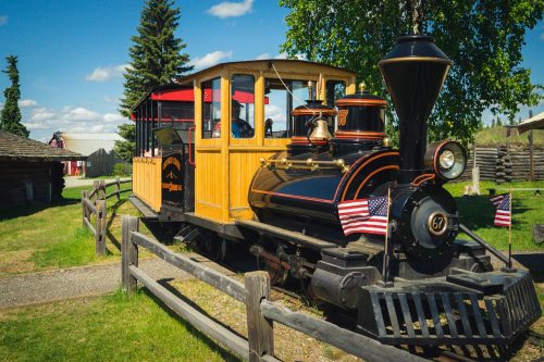 A vintage steam train with wooden features, flags, and a scenic background of trees and blue sky.
