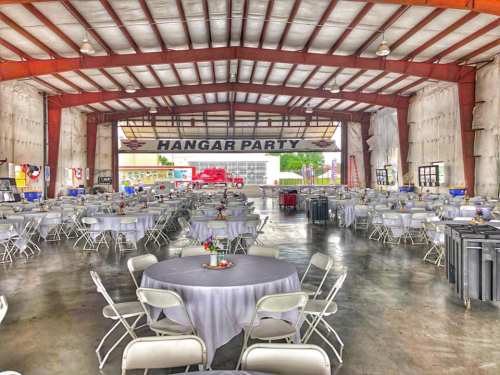 A spacious event hall with round tables and chairs set up for a party, featuring a "HANGAR PARTY" banner.
