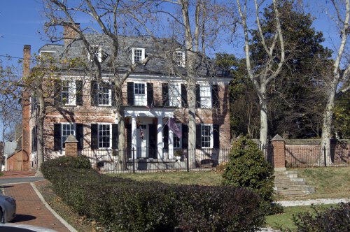 A large brick house with a slate roof, surrounded by trees and a manicured lawn, featuring a front porch and iron fence.