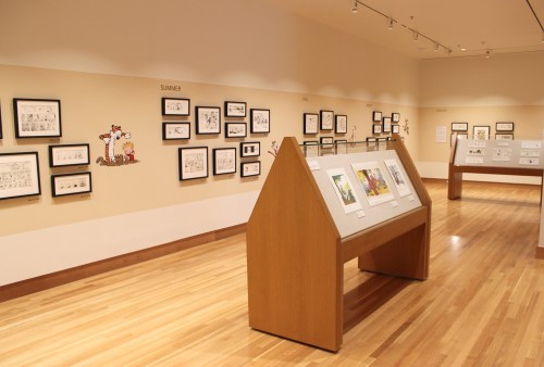 Art gallery interior featuring framed artworks on walls and display tables with illustrations. Warm wooden flooring.