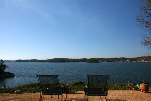 Two black chairs facing a calm lake under a clear blue sky, surrounded by greenery and distant hills.