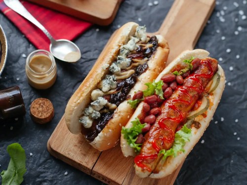Two gourmet hot dogs on a wooden board: one with blue cheese and mushrooms, the other with chili, beans, and lettuce.