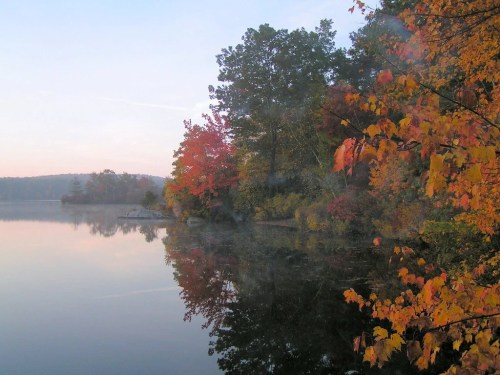 A serene lake at dawn, surrounded by vibrant autumn foliage reflecting on the water's surface.