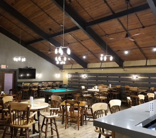 Interior of a rustic dining area with wooden tables, chairs, and a pool table under a high, beamed ceiling.