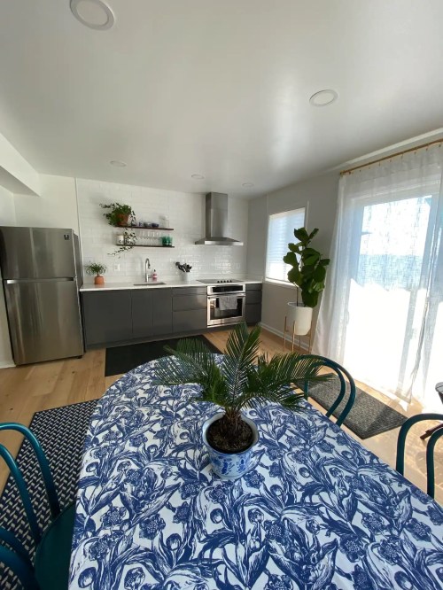 A modern kitchen with a blue patterned tablecloth, potted plant, stainless steel appliances, and bright natural light.