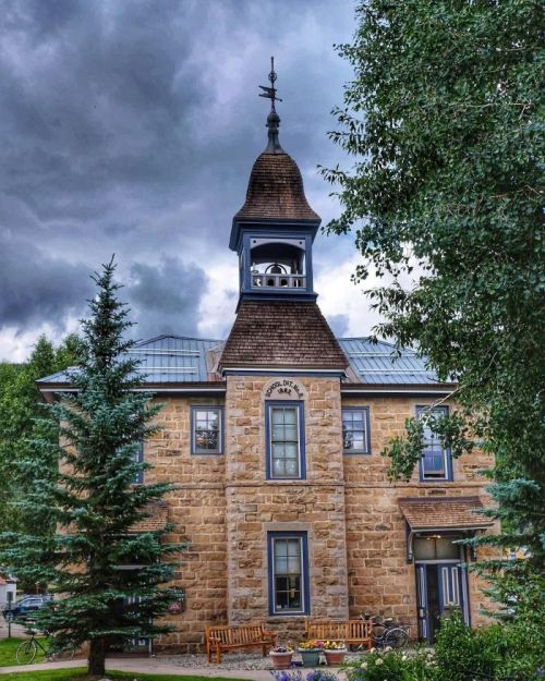 Historic stone building with a clock tower, surrounded by trees and a cloudy sky.