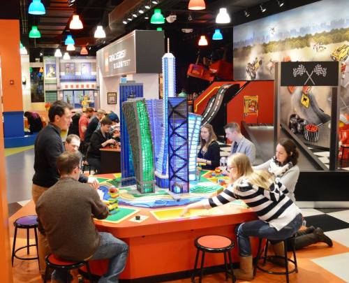 A group of people gathers around a colorful model city at a playful indoor attraction, engaged in a game.