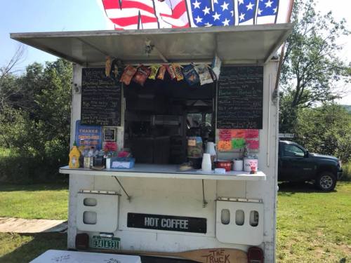 A food truck with a menu board, serving hot coffee and snacks, set in a grassy area with American flags above.